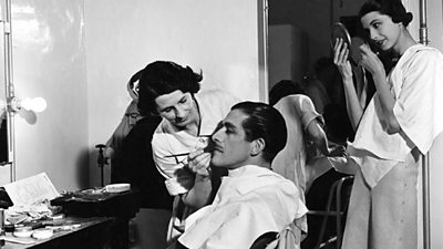 A man is sitting down with a cloth around his suit while a woman paints black makeup on his face.