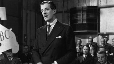 A man in a sharp double-breasted suit and pencil moustache stands in front of a camera