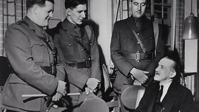 Three men in officer's uniforms stand speaking to a seated man in a suit. 