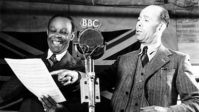 A man points at a script while another man laughs. In front is a ý microphone and behind a large Union Flag. 