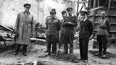 A group of officers stand in the ruins of a building. Richard Dimbleby is in a belted raincoat on the left. 