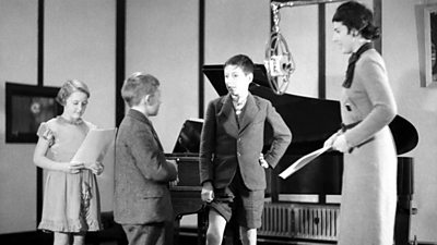 Three children reading a script by a large microphone, supervised by a women. A grand piano is in the background. 