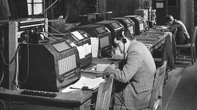 A long desk of transcribers wearing headphones listening and writing down what they hear on the radio.