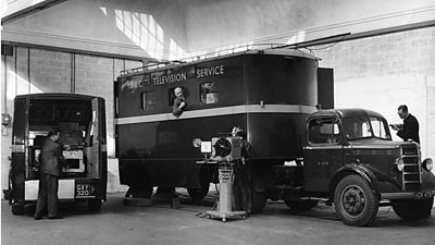 Two Bedford trucks with ý Television Service on the side. Various male crew are working by the vehicles and a large camera on a tripod is in the centre.