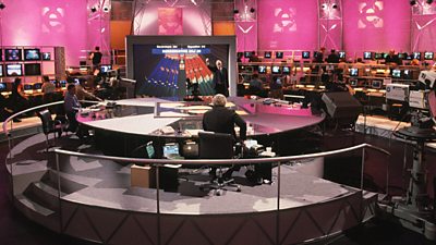 The election studio with a bright pink hue in 1997, the presenters at a giant round desk with the rest of the studio orbiting them. There are hundreds of monitors. 