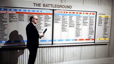 Robert McKenzie holding a clipboard in front of a graphic of constituencies titled 'The Battleground'.