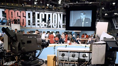 A behind the scenes shot of the 1966 election studio - a large TV camera facing the presenters, with a bank of monitors and a large number of people in the background. 