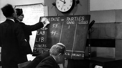 A production member rubs off numbers on a chalkboard with 'State of Parties' written on it with gains and losses tallied next to CON, LAB, LIB and OTHERS.