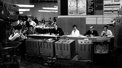 Richard Dimbleby, centre, surrounded by presenters at the main desk, with a bank of monitors facing him. The swingometer graphic is on the wall behind and a large TV camera pointing at them.  