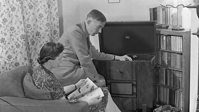 A husband and wife in their chintzy 1950s living room gathered around a large walnut radio. The man is fiddling with the dial. 