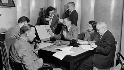 A large group of journalists around a desk examining the results on paper. Two have showerhead-style commentating microphones. 