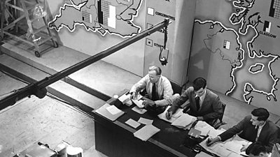 A overhead shot of the presenters sat at their election desk in the studio, a large UK map on the wall behind and a microphone boom visible. 