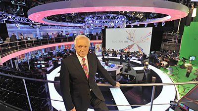 David Dimbleby posing on the balcony over the giant circular election studio. The formality has returned. There is now a giant green screen area at the side of the studio. 