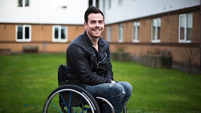 A young man in a wheelchair. He is wearing a leather jacket and ripped jeans.