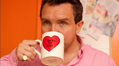 A man sips a mug with 'you're my hero'. He's wearing a lot of gold jewellery