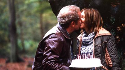 A man holding a birthday cake kisses a woman in a forest.