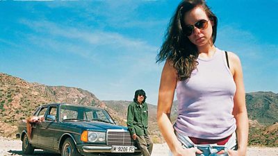 A woman with her hands in pockets looks down in the midday sun, her car behind with a louche man resting on the bonnet. 