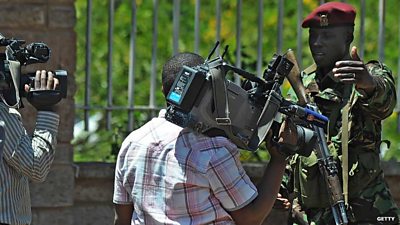 Men each carrying a video camera are speaking to soldier who has his arm raised, the soldier is wearing camouflage clothing and a red hat