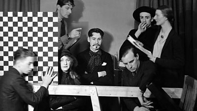 A group of actors waiting to go on air while the producer prompts them from a script - the central actor sits folded armed has a rather fake-looking moustache and beard. A stage hand is sliding across a chess-board like chequerboard as a scene divider.