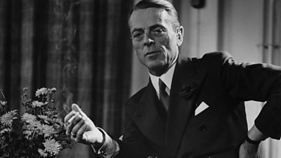 A rather severe-looking man in a suit smoking a cigarette in his office. He has one hand on his waist.