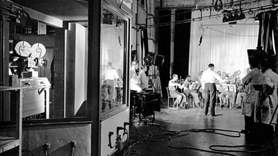 A soundproof glass booth with a film camera pointing at an orchestra playing some way off in another part of the studio. 