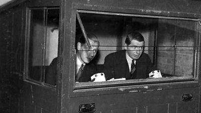 Two male commentators in a shed-like commentary box with a large glass window