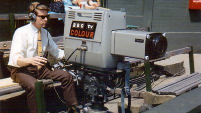 A cameraman in shirt and tie operates a large grey camera.