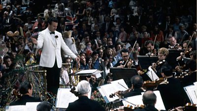 A conductor in a white jacket conducts the orchestra at the Last Night of the Proms. The photo is in colour. 