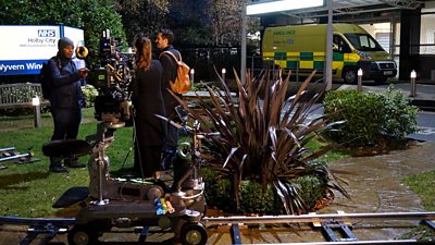 A camera on a track is in the foreground as a director talks to actors outside the external set of Holby City. An ambulance is in the background