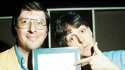 Paul McCartney pulls a silly face pointing at a frame held by Simon Bates