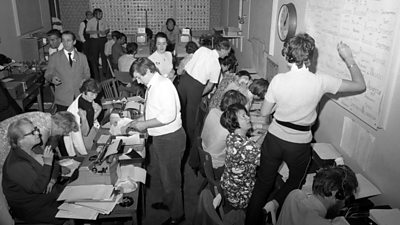 A busy newsroom - people writing on whiteboards while other sit at typewriters