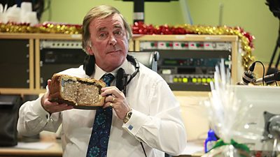 Terry Wogan looking bemused in a Christmassy studio holding an enormous pork pie. 