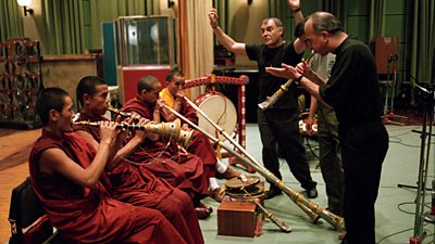 Four Tibetan monks in traditional dress play instruments, while Western musicians join in.  