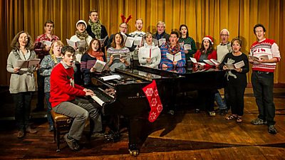 The ý Singers, all wearing Christmas jumpers, sing around a Steinway grand piano