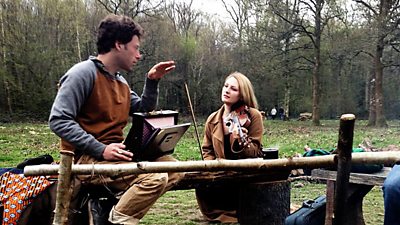 A female cellist in woodland. A sound recordist is making last minute checks. 