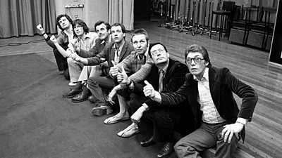 The cast of the radio Hitchhiker’s Guide to the Galaxy pose on a bench as if thumbing a lift