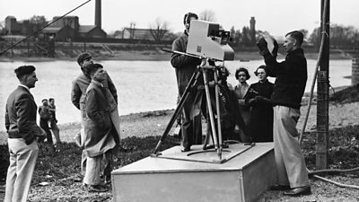 A large early EMI camera on a tripod and raised platform on the shore of the Thames. Engineers are doing tests on the equipment.