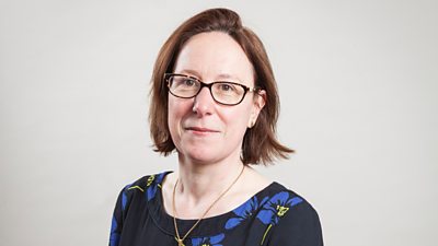 Clare Sumner posing for her profile photo with a slightly amused smile. She wears glasses, a primrose pattern top and a gold necklace.
