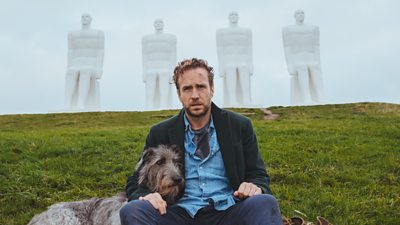 Rafe Spall looks dishevelled sitting next to a Danish statue, his arm around an equally sad looking Irish Wolfhound 