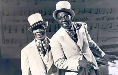 Two black singers in white coats, spotted cravats and top hats.