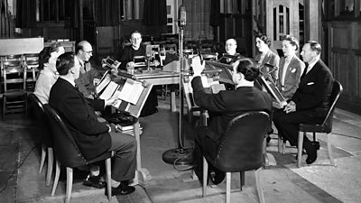 A group of men and women congregated around a microphone in a church, each with a lectern stand. A vicar sits at the head. 