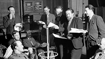 A large group of relaxed men gathered around a microphone in a studio, scripts in hand.