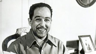 Langston Hughes looks cheerfully at the camera, a photograph in a frame at his side.