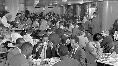 Men and women of all different nationalities eat in a very crowded canteen. 