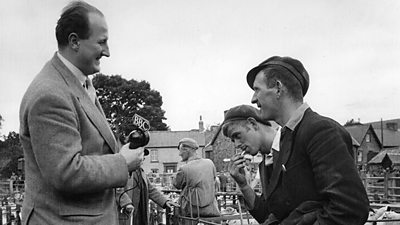 A presenter in a tweed jacket interviews a farmer in a flat cap using a 'showerhead' type microphone. Various sheep pens can be seen behind. 