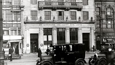 The front of a stone-faced building. A 1920s taxi waits in a rank on the street below. 