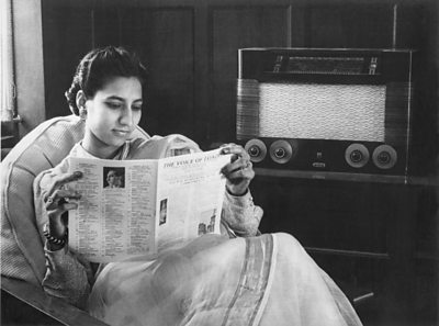 An Indian woman in a sari reads a magazine 'the voice of london' while listening to a large wireless radio. 