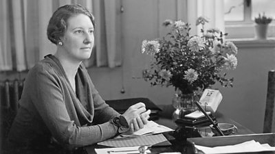 A woman sits at a desk, looking wistfully into the distance. A large bunch of flowers is on her desk.