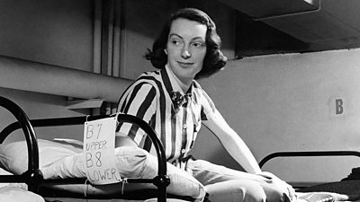 A woman with a quizzical smile wearing a stripy shirt and trousers sits on the top of a bunk bed. 