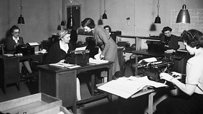 Women typists sit behind five desks. A supervisor talks to the women in the middle